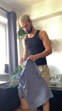 Muž s krátkými blond vlasy a plnovousem skládá světle modrou košili v místnosti s rostlinami, oknem a vintage šicím strojem.