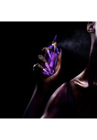 Flacon de parfum en verre violet avec un accent doré, tenu par une main aux ongles longs et pointus, émettant une fine brume sur un fond noir.