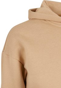 Beige kapucnis pulóver puha textúrával, keresztszegéssel ellátott kapucnival, süllyesztett vállakkal és bordás részletekkel a mandzsettákon és az alján.