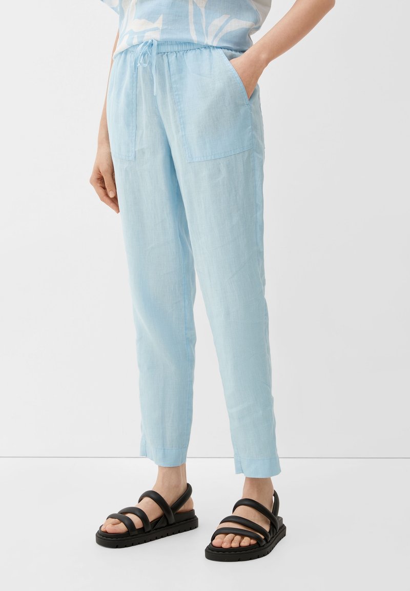 Lichtblauwe linnenbroek met een elastische tailleband, voorzien van twee voorkruiken en taps toelopende pijpen, gecombineerd met zwarte sandalen met bandjes.