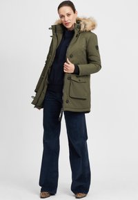 Olive green parka jacket with a fur-lined hood, button and zipper closure, and multiple pockets. Worn over a dark sweater and navy corduroy pants.