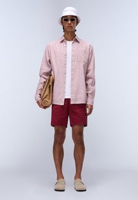 Gestreepte roze en witte button-up shirt met zak, rode shorts, beige instappers, witte bucket hat en een bruine tas in de linkerhand.