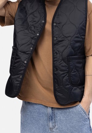 Person trägt ein braunes T-Shirt, helle blaue Jeans und eine offene schwarze gesteppte Weste mit Druckknöpfen und Vordertaschen.