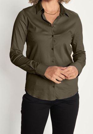 Frau trägt eine olivgrüne Bluse mit Knöpfen und langen Ärmeln sowie schwarze Hosen, die Hände auf Taillenhöhe verschränkt, vor einem einfarbigen Hintergrund.