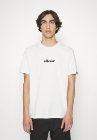 Weißes Baumwoll-T-Shirt mit einem schwarzen Logo mit der Aufschrift "ellesse" auf der Vorderseite. Kurze Ärmel und runder Ausschnitt. Schlichtes und klassisches Design.