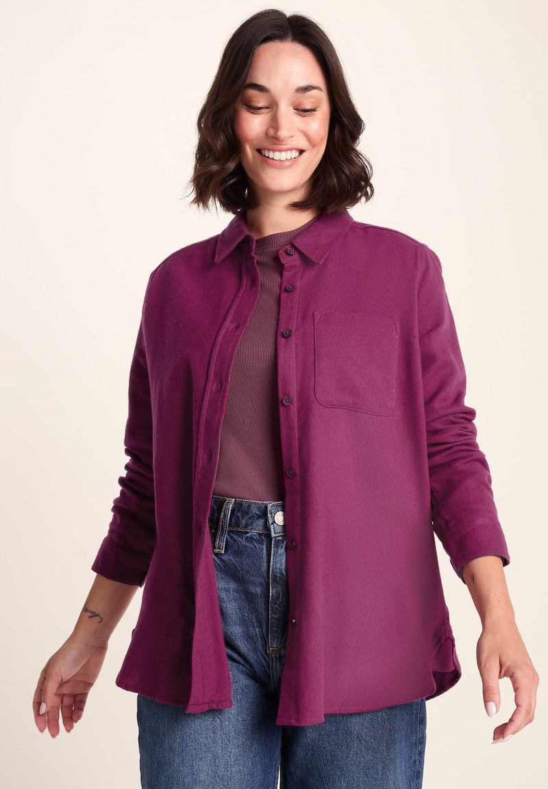 Purple button-up shirt with a front pocket, made of a soft material; worn over a brown top, paired with high-waisted denim jeans.