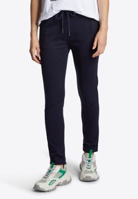 Marinblå joggers med dragsko i midjan, muddar vid anklarna och en slät tygtextur, som är kombinerade med kraftiga vita och gröna sneakers.