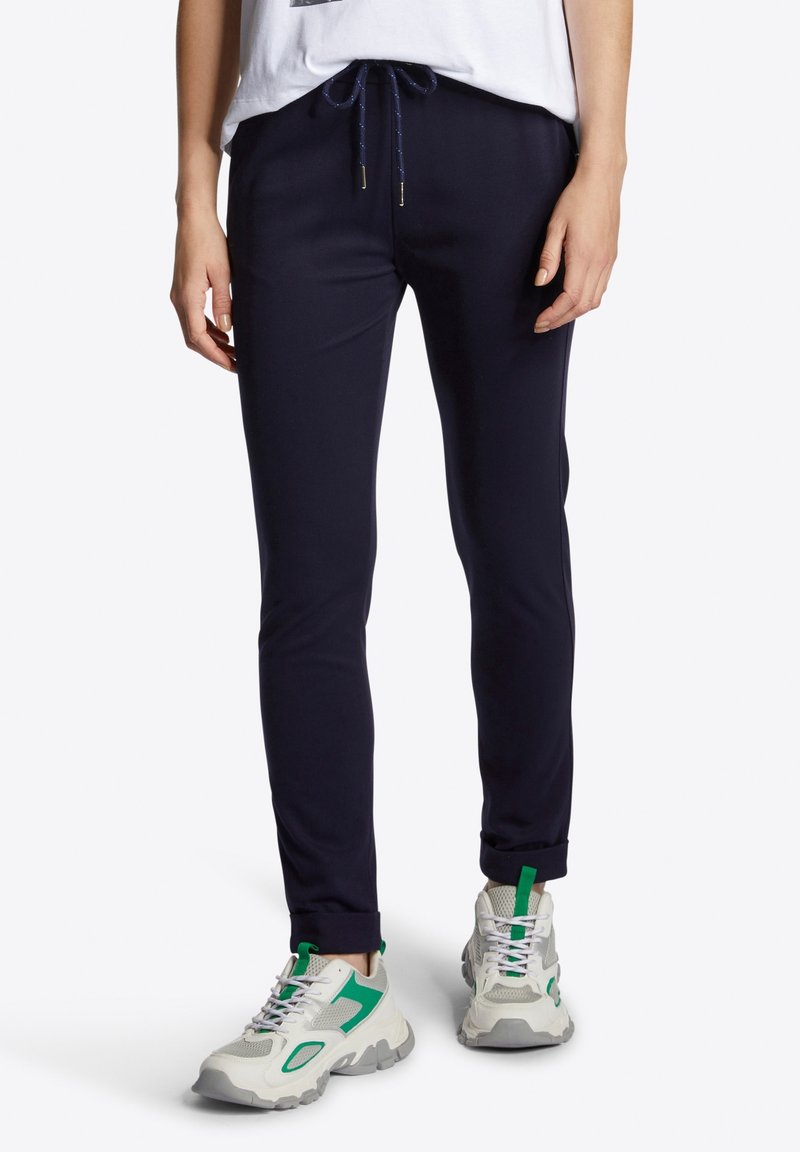 Marinblå joggers med dragsko i midjan, muddar vid anklarna och en slät tygtextur, som är kombinerade med kraftiga vita och gröna sneakers.