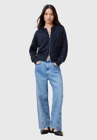 Perfekt für den Alltag: Ein marineblauer, flauschiger Reißverschluss-Cardigan mit rundem Ausschnitt, kombiniert mit hellblauen hochgeschnittenen, weit geschnittenen Jeans und schwarzen Ballerinas.