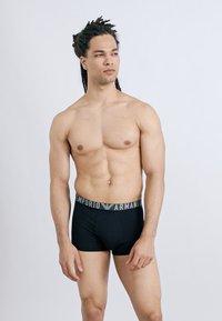 Άνδρας χωρίς πουκάμισο με ντραντλόκ, που φοράει μαύρα μποξεράκια Emporio Armani, στέκεται μπροστά σε μονόχρωμο ανοιχτό γκρι φόντο, κοιτάζοντας προς τα πλάγια.