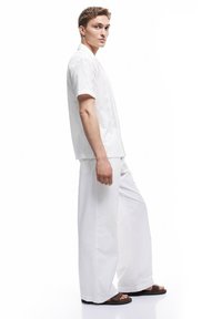 Weißes Kurzarmhemd mit einem strukturierten Muster und einem Button-Down-Kragen, kombiniert mit lockeren weißen Hosen und braunen Sandalen.