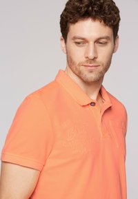 Polo de manga corta en algodón naranja vibrante con un acabado texturizado, que presenta una tapeta de tres botones y un logo bordado en el pecho.