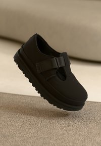 Chaussure noire style Mary Jane avec une large bride et une fermeture velcro, semelle épaisse, présentée sur un fond neutre et doux.