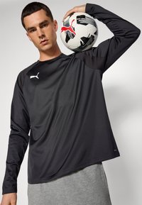 Svart langermet treningsskjorte med teksturert mønster, med en hvit Puma-logo, kombinert med grå joggebukser og en svart-hvitt fotball.