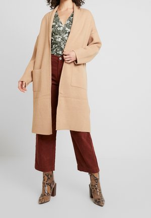 Femme portant un long cardigan beige ouvert sur le devant, un haut fleuri vert, un pantalon large couleur rouille et des bottines à talons imprimé peau de serpent.