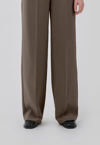 Person wearing wide-leg brown pants long enough to cover tops of black square-toe shoes, standing against plain background.