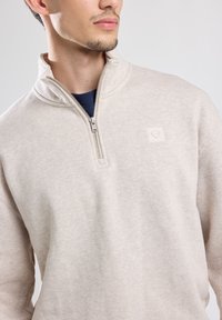 Pullover beige chiaro con colletto alto, zip frontale a metà e piccolo logo testurizzato sul petto. Tessuto morbido e testurizzato.