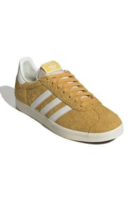 Semišová žlutá teniska s třemi bílými pruhy, texturovaným povrchem, plochými béžovými tkaničkami a bílou gumovou podrážkou. Logo viditelné na jazyku boty.