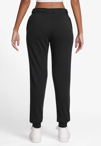 Kvinne iført svarte joggebukser og hvite sneakers, vist fra bakgrunnen mot en enkel hvit bakgrunn.