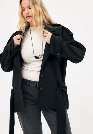 Kvinde justerer sort oversized blazer over cremefarvet top, iført sort halskæde og falmede sorte jeans mod en ensfarvet hvid baggrund.