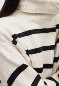 Pull en tricot beige avec un col montant, affichant des rayures noires horizontales. Texture douce et coupe ample avec des épaules dénudées.