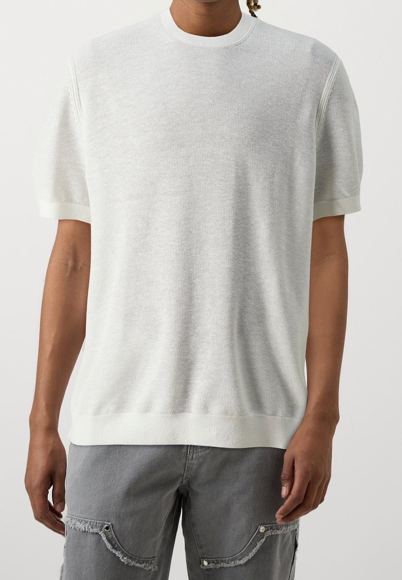 Personne portant un pull en maille blanc à manches courtes et un pantalon gris usé, debout devant un fond clair uni.