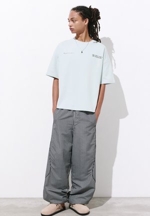 Personne avec des dreadlocks portant un t-shirt blanc ample avec un texte et un pantalon large à carreaux noir et blanc, debout devant un fond blanc.