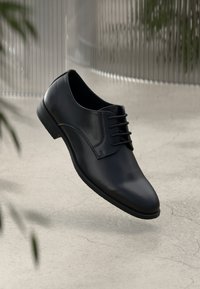 Pantof cu toc din piele neagră, cu șireturi, prezentat elevat pe un podea neutră texturată și o suprafață reflectoare verticală neclară.