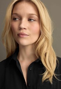 Schwarzes Hemd mit Kragen und Knopfdetails, getragen von einer Person mit welligem blonden Haar. Sanftes Licht hebt die Gesichtszüge und die Hautstruktur hervor.