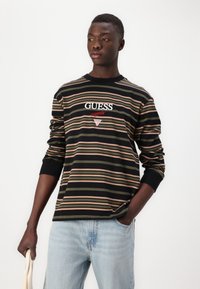Svart langermet stripete skjorte med grønne og rosa horisontale striper, GUESS-logo i hvit og rød brodering, brukt sammen med lyseblå jeans.