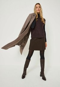 Brun uldtrøje lagdelt over en sort turtleneck, parret med en mørkebrun nederdel og knæhøje brune læderstøvler. En grå frakke hænger over den ene arm.