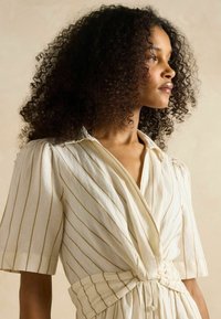 Gaišas krāsas svītrainā kleita ar apvārsni, īsām piedurknēm un mezgla jostas detaļu. Izgatavota no struktūras auduma.