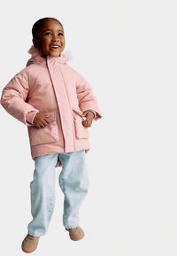 Roze gewatteerd jack met capuchon, met twee voorzakken en geribbelde manchetten, gedragen over lichtblauwe spijkerbroek en bruine schoenen.