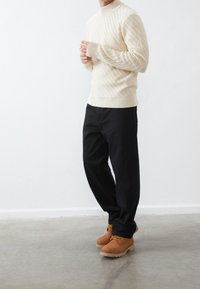 Man som bär en krämfärgad ribbad tröja, svarta byxor och tan-färgade arbetsskor, stående på en betonggolv mot en vit vägg.