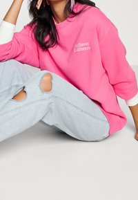 Kvinna sittande på golvet iförd en ljusrosa Hollister California-tröja, ljusblå slitna jeans och långt mörkt hår med rosa örhängen.