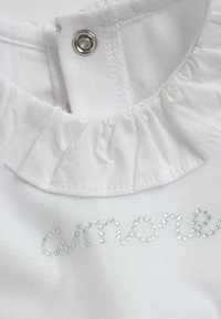 Vêtement blanc avec un col volanté et une fermeture à pression. Présente un texte en sequins argentés épelant "amore" sur un tissu texturé.