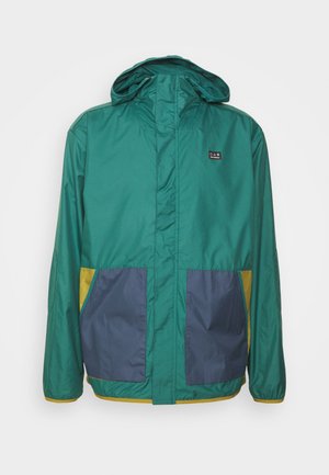 Zöld, könnyű dzseki kapucnival, kontrasztos tengerészkék és sárga zsebekkel, sima textúrával és elöl zippzárral.