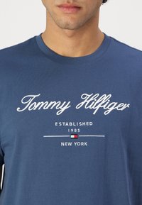 Blå bomull t-shirt med vit broderad text: "Tommy Hilfiger," "ETABLERT 1985," och "NEW YORK," med en liten logotyp som accent.