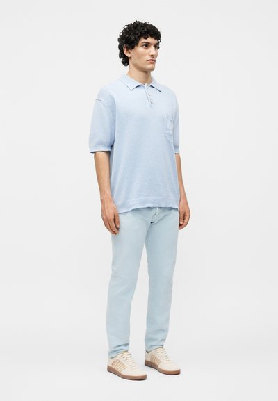 Άνδρας με σγουρά μαλλιά που φοράει ανοιχτό μπλε πόλο με κοντά μανίκια, ανοιχτό μπλε τζιν και μπεζ αθλητικά παπούτσια, στέκεται μπροστά σε απλό φόντο.