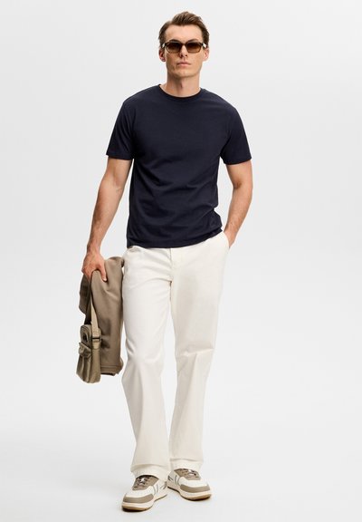 Άνδρας που φοράει σκούρα γυαλιά ηλίου, μαύρο μπλουζάκι, λευκό παντελόνι, μπεζ αθλητικά παπούτσια, κρατώντας μια μπεζ ζακέτα και τσάντα ενώ περπατά προς τα εμπρός σε λευκό φόντο.