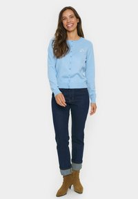 Világoskék kardigán, kerek nyakkal, gombos záródással és kis szív hímzéssel. Sötét farmernadrággal és világosbarna hegyes orrú csizmával párosítva.