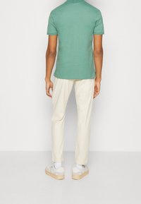 Polo vert, coupe slim, tissu texturé ; associé à un pantalon crème. Des baskets blanches avec des semelles beiges complètent le look.