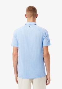 Ljusblå kortärmad pikétröja med ett strukturerat mönster, kontrasterande krage och ärmdekorationer samt en logga på övre ryggen.