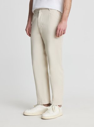 Uomo con pantaloni beige a gamba dritta e scarpe da ginnastica bianche, in piedi davanti a uno sfondo chiaro e semplice.