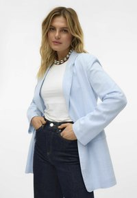 Blazer bleu clair avec une finition texturée, fermeture à un bouton et manches larges, associé à une chemise blanche et un jean en denim foncé.