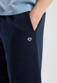 Tummansiniset puuvillaiset verkkaripantsit, joissa on sivutasku ja pieni punaisesta ja sinisestä brodeerattu logo vyötärön läheisyydessä. Pehmeä rakenne ja istuva malli.