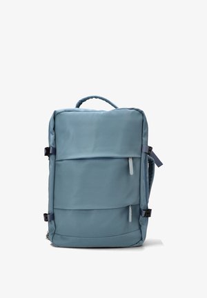 Mochila rectangular de color azul claro con asa superior, dos bolsillos frontales con cremallera y hebillas laterales ajustables, colocada de pie sobre un fondo blanco.