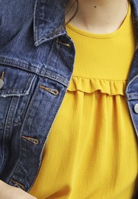 Giubbotto in denim con accessori in argento sopra un top giallo caratterizzato da un tessuto testurizzato e un colletto arricciato. Primo piano dei capi.