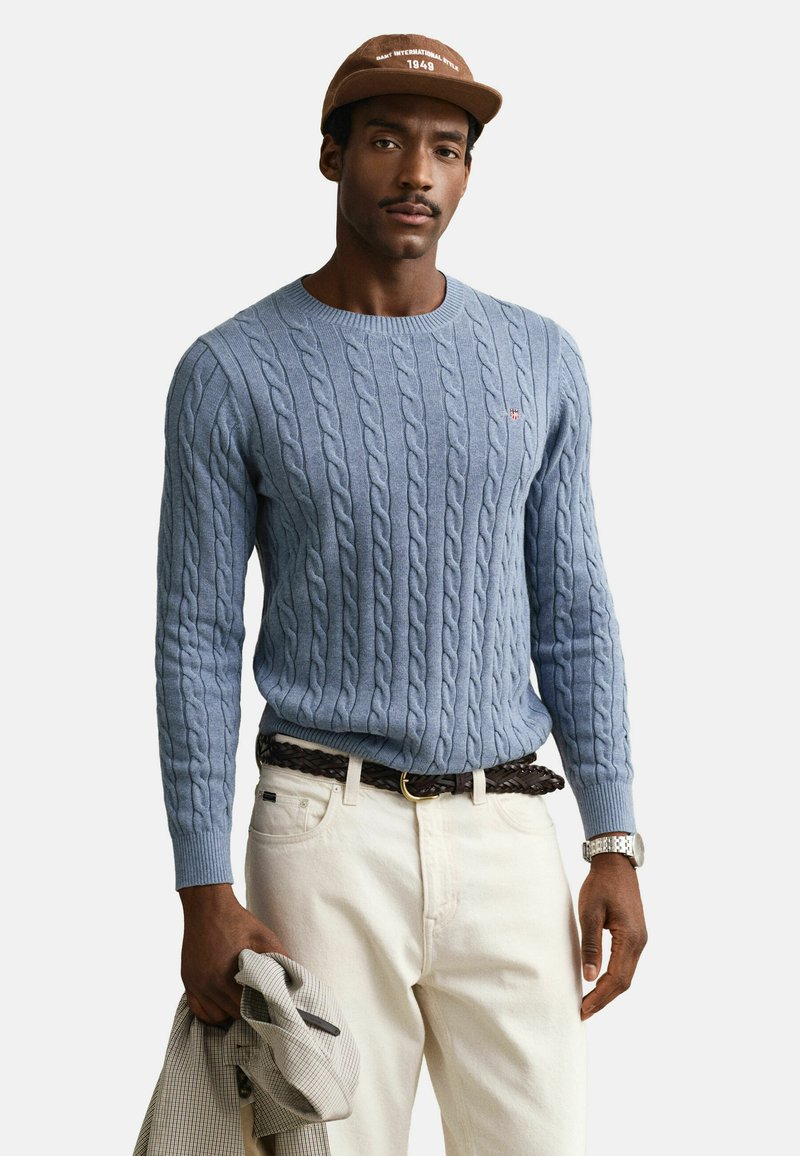 Homme portant un pull bleu clair en maille torsadée, pantalon crème avec ceinture tressée marron, casquette marron, montre-bracelet argentée, tenant une veste beige à carreaux.