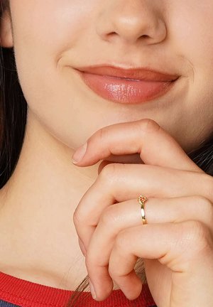 Gold ring with a small heart-shaped design atop, worn on a finger, with a hand resting near a smiling face featuring glossy lips.